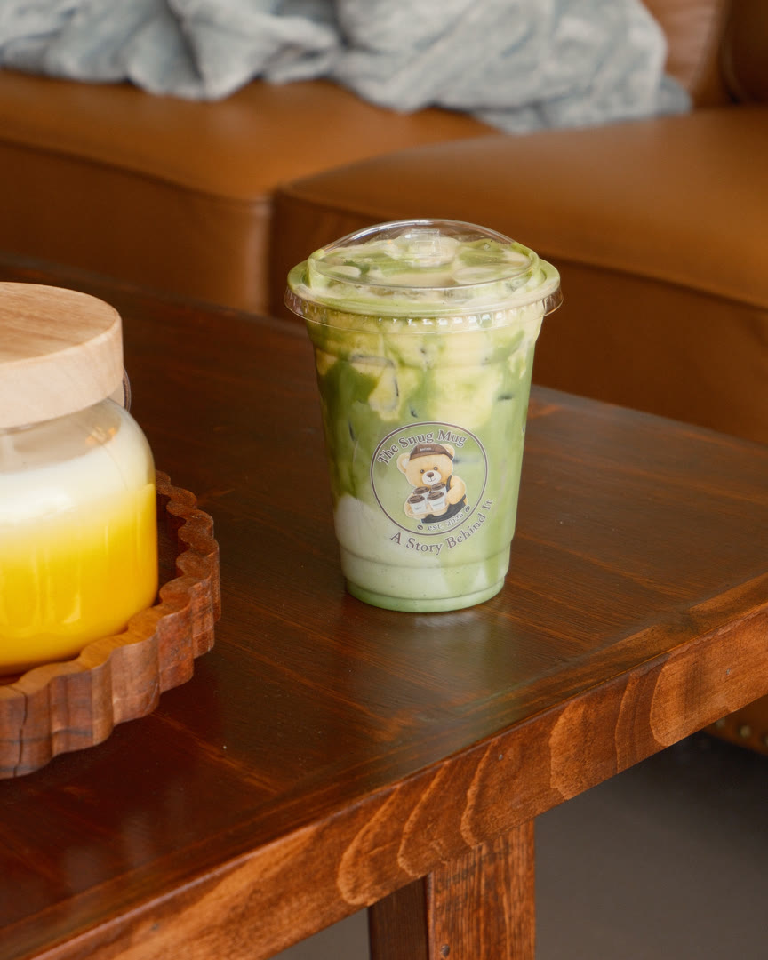 An iced matcha in The Snug Mug's own printed cup, on a walnut table